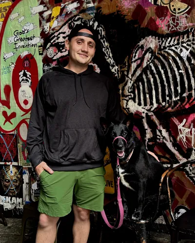 John and Kona, a black dog on a pink leash, in front of a wall covered in stickers and street art